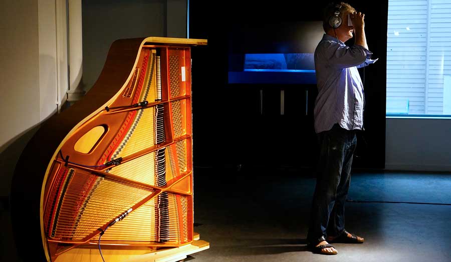 Man in headphones standing in the dark next to a grand piano laying on it's side and lit up inside